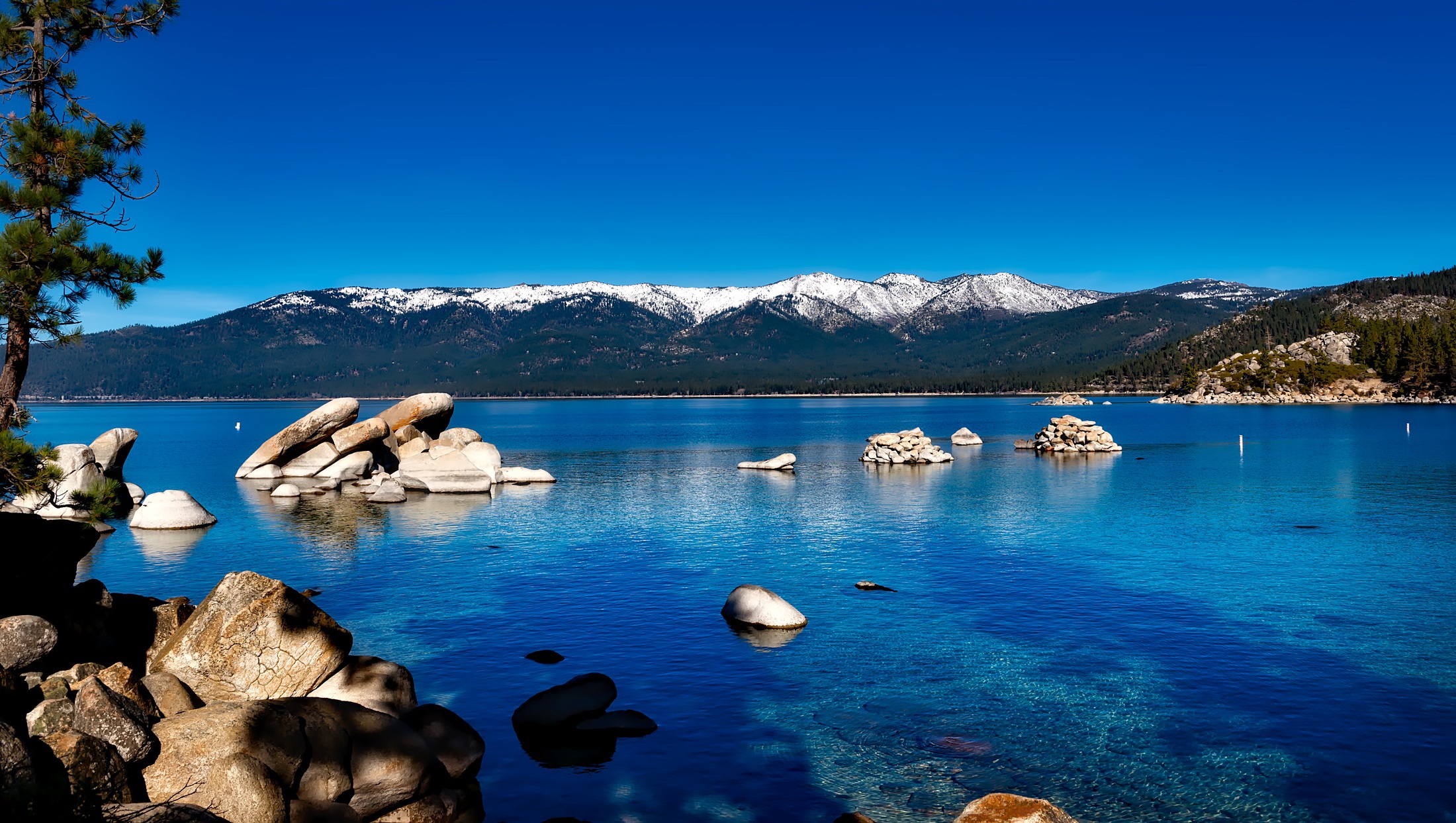 Lake Tahoe's clear blue waters, rocks in the foreground and snow on the distant mountain peaks