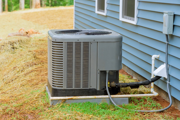 Photo of an air conditioner unit outside a house on a sunny day, surrounded by dry grass.