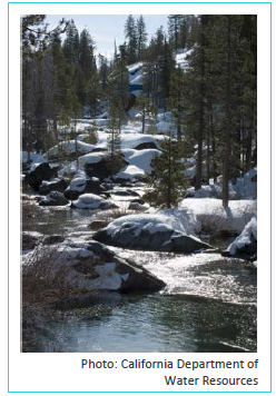 Photograph of a mountain stream in the Sierra Nevadas surrounded by trees and snow covered rocks. It is a clear sunny day and you can see the snow melting in watery streaks down the rocks and into the stream.